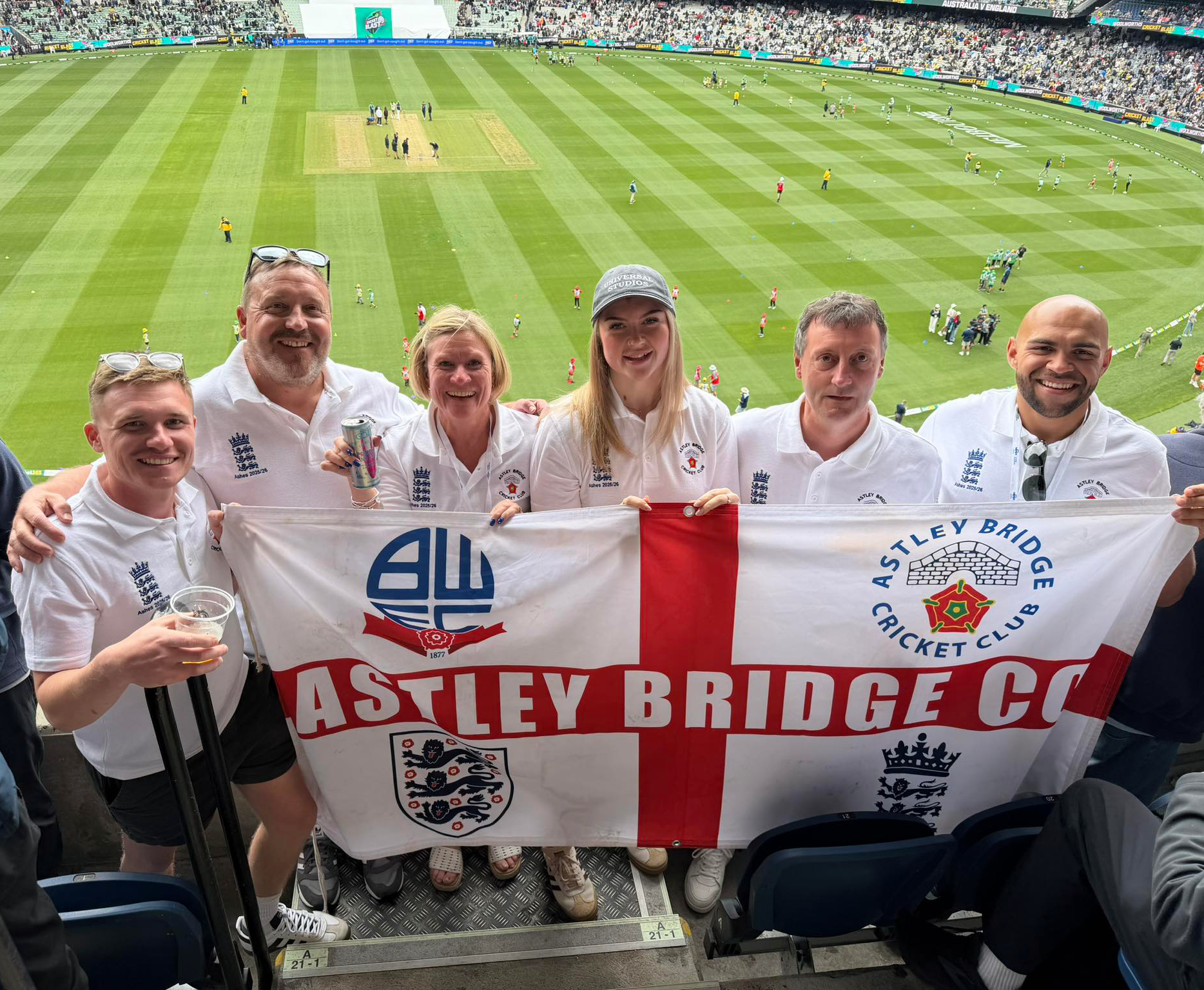 A group photo of a few of Astley Bridge Cricket Club's Members holding a large flag with logos of different sports teams. 
